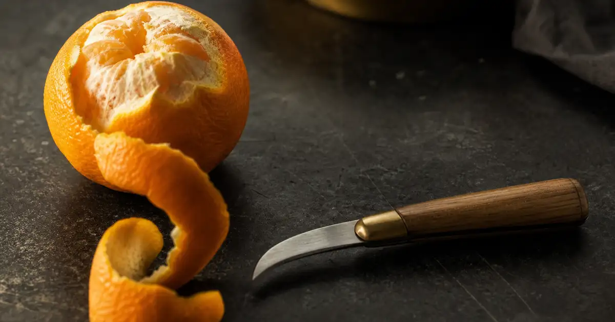 A curved bird's beak paring knife beside a partially peeled orange