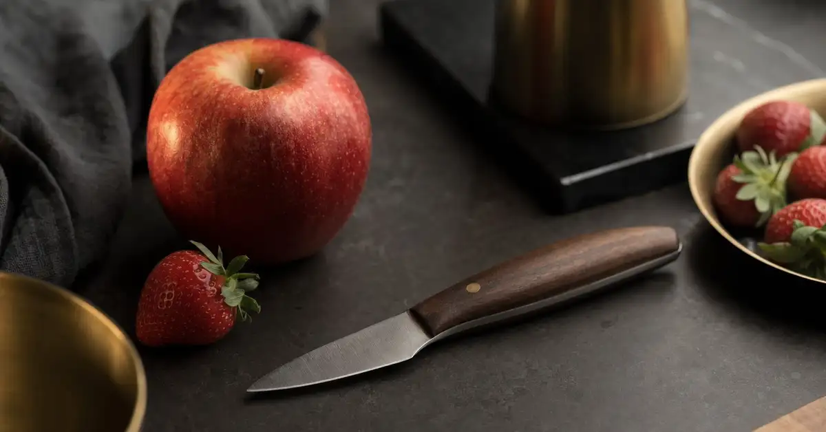 A small paring knife beside an apple and a strawberry on a matte slate counter in warm light